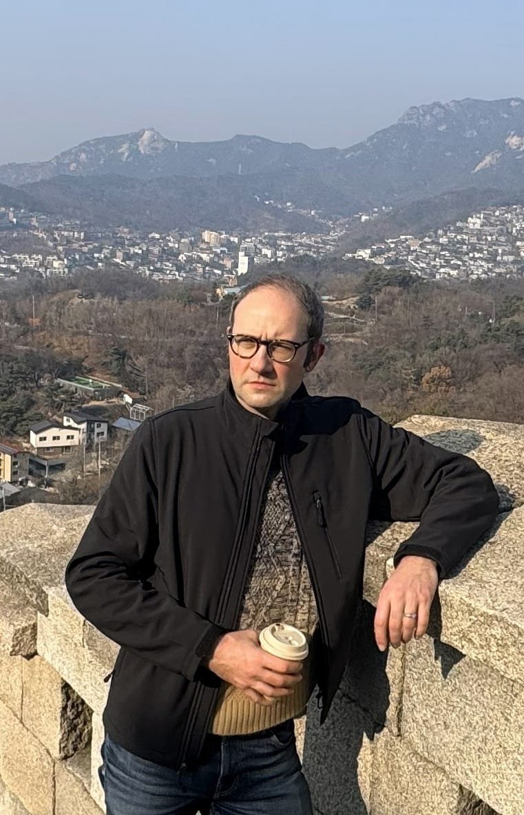 A photo of CMO Jesse Swingle, a man in a jacket, glasses and sweater, standing on a wall in Seoul, discussing entering new markets with a coffee in one hand.