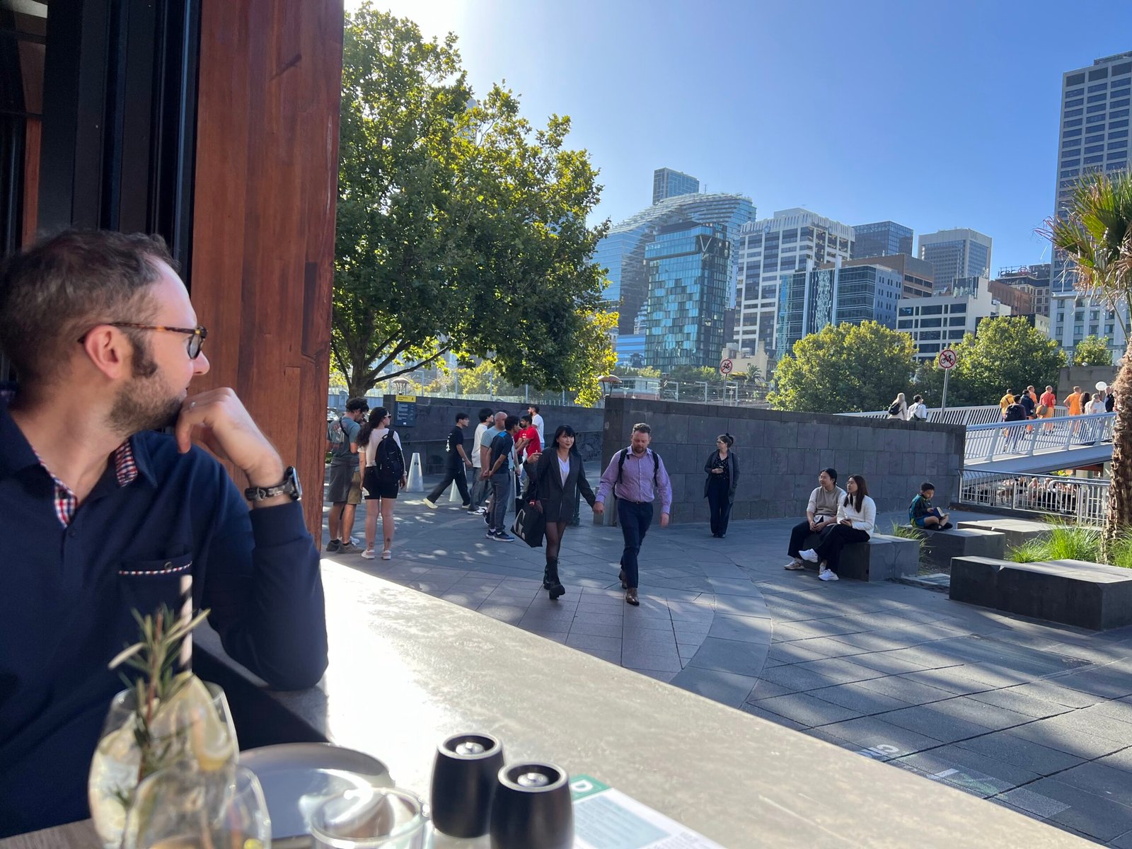 Jesse seated at an outdoor restaurant table, looking out toward a busy waterfront promenade and city skyline, appearing thoughtful as people walk past in the background.