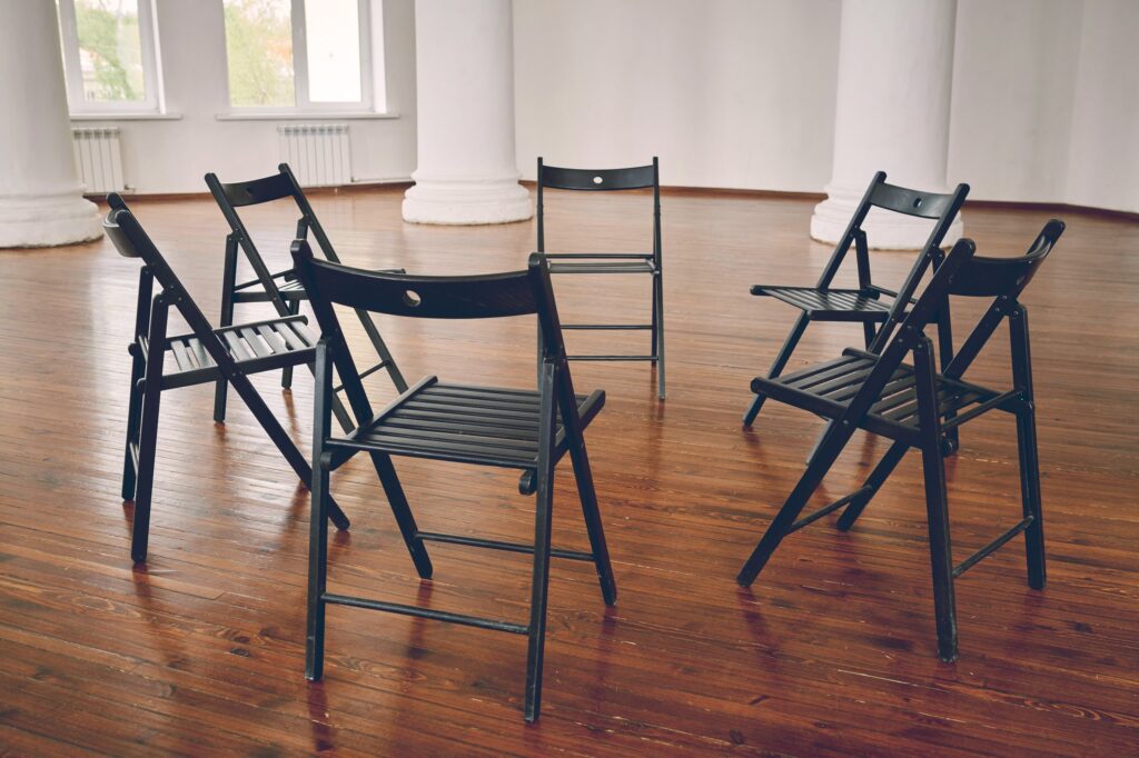 Black folding chairs set loosely in a circle, facing different directions, on a wooden floor in an empty hall