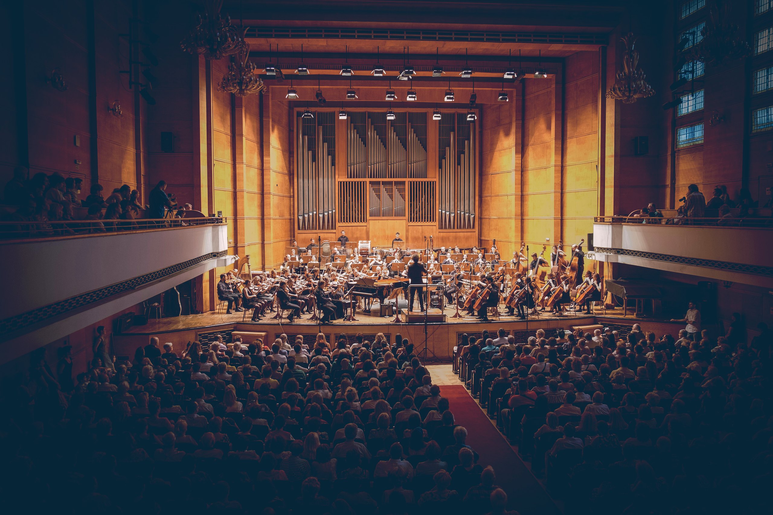 An orchestra performing on stage in a wood-panelled concert hall, seen from behind a full audience